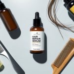A bottle of hair repair serum surrounded by various hair care products on a clean, white surface. A comb and hairbrush are nearby, with strands of hair scattered around